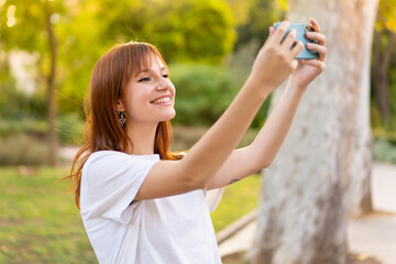 Obraz premium Young pretty redhead woman at outdoors using mobile phone and doing a selfie