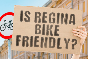The question " Is Regina bike friendly? " on a banner in men's hand with blurred background. Transportation. Zero waste. Bicycle lane. Streets. City. Safety. Insecure. Road signs. Dangerous