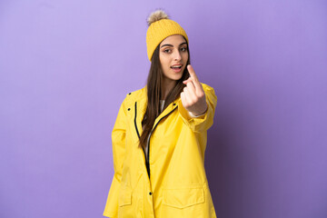 Young caucasian woman wearing a rainproof coat isolated on purple background doing coming gesture