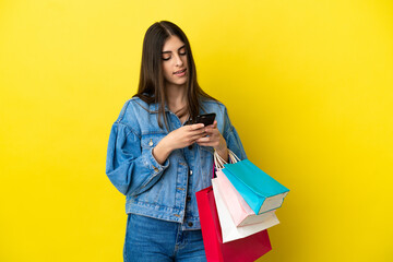 Young caucasian woman isolated on blue background holding shopping bags and writing a message with her cell phone to a friend