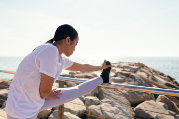 young asian sportswoman stretching legs by the sea, concept of sport and active lifestyle