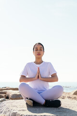 Naklejka na ściany i meble vertical photo of a young asian woman relaxed meditating sitting outdoors with hands together and eyes closed, concept of spirituality and relax