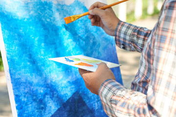 Close-uo of unrecognizable man using color palette while painting background on easel outdoors