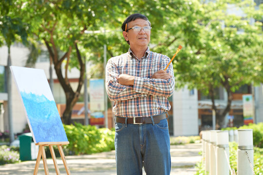 Smiling Mature Asian Artist In Eyeglasses Wearing Pencil Behind Ear Standing With Crossed Arms Outdoors While Finding Inspiration