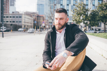 Obraz premium Young adult caucasian bearded man outdoors sitting waiting holding smartphone