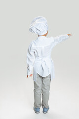 Back view of little boy in white cook uniform and huge chef's hat standing isolated on white studio background.