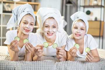 Portrait of happy mature woman lying on sofa with her pretty teen daughters and doing vitamin mask on face. Beauty procedures for healthy and smooth skin.