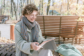 Fototapeta premium pensive teenager girl in a knitted hat with a book and a yellow autumn leaf in her hands sits near a tree in the park