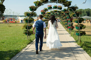 The bride and groom walk down the road to the wedding day, the view from the back. A man in an elegant suit of blue tones, with a vest. A woman in a white wedding dress.