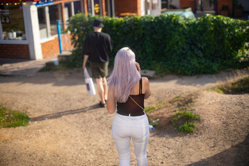 People walk along a sandy road with interspersed greenery in the park. A girl with purple hair, in white pants and a black T-shirt, a view from behind. Sunny day.
