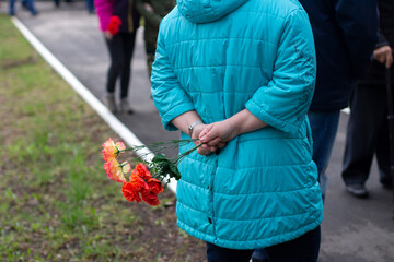 A woman in a blue jacket stands with flowers behind her back. View from the back. The street of the city, the crowd of people. Victory Day.
