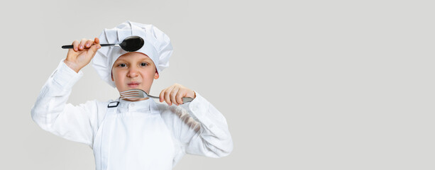 Close-up portrait of little happy boy in white cook uniform and huge chef's hat with big spoon and fork isolated on white studio background.