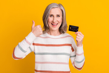 Photo of adorable shiny retired woman dressed white sweater holding debit card showing thumb up isolated yellow color background