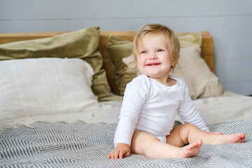 A small child with blond hair and white clothes sits on the bed and smiles