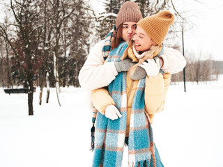 Two young beautiful smiling hipster female in trendy warm clothes and scarfs.Carefree women posing in the street in park. Positive pure models having fun in snow. Enjoying winter moments. Xmas concept