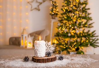Selective focus on snowball pattern candle burning, decorated with pine tree wood disc, white snowflake decoration and fake snow on table. Blurred Christmas home living room on background.