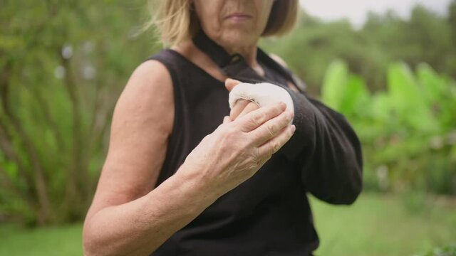 Senior elderly woman massaging hand suffering from joint pain. Older woman with injury broken arm in cast outside in summer. Old lady touching wrist in gypsum sling bandage feeling hurt. 