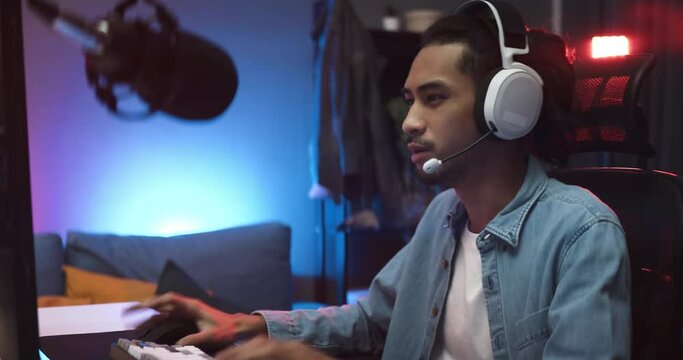 Young Asian man playing online computer video game, colorful lighting broadcast streaming live at home. Ecstatic celebration winning a match. Gamer lifestyle, E-Sport online gaming technology concept