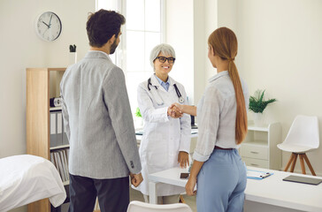 Congratulations, you're gonna have a baby. Happy patients exchange handshake with smiling senior family doctor. Young married couple planning pregnancy and seeing OB GYN or IVF specialist together