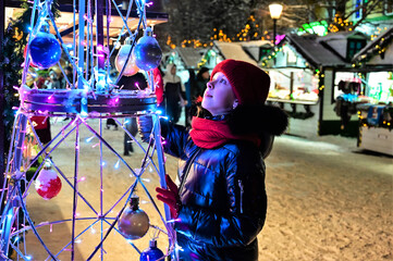 Fototapeta na wymiar beautiful girl on street is looking at creative Christmas tree made of metal, bright garlands and decorations. New Year's festive mood. Christmas tree design