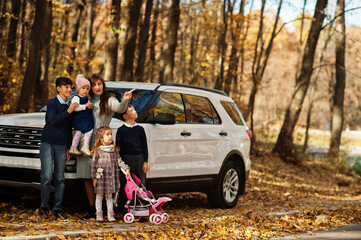Mother stand near white suv car with four kids in autumn park. Family walk in fall forest. - obrazy, fototapety, plakaty