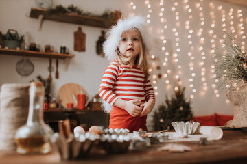 little girl in a Santa hat and red striped pajamas cooks a Christmas cake in a beautiful kitchen decorated with a garland. merry Christmas and happy New Year and Holidays. hygge. High quality photo