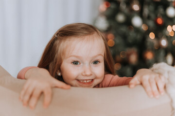 cute little blonde girl in pink clothes is kneeling in a chair against the background of a Christmas tree. child is waiting for the advent of Christmas and new year. winter concept. High quality photo
