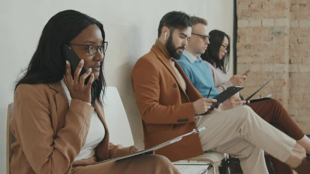 PAN shot of cheerful young African-American woman in glasses and formal wear sitting on chair in queue for job interview and talking on mobile phone
