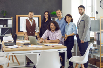 Team of happy creative business professionals at work. Group portrait of company workers sitting or standing by the table during meeting in modern office interior. Concept of success and teamwork