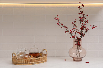 Hawthorn branches with red berries in vase and aromatic tea on white table indoors