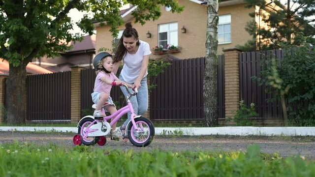 A young mother teaches her daughter to ride a bike. Little girl in helmet learns to ride a pink bike under the supervision of mom
