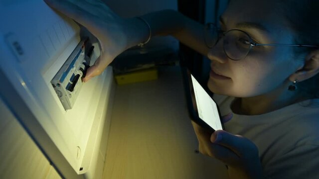 Young woman with a phone checks a row of circuit breakers in fuse box during a power outage in an area
