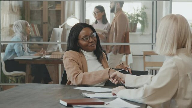 Tracking shot of cheerful African-American woman in glasses singing document and shaking hands with female HR manager after successful meeting or job interview