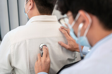 Male doctor in medical face mask using stethoscope to listening heart and lungs from male patient's...