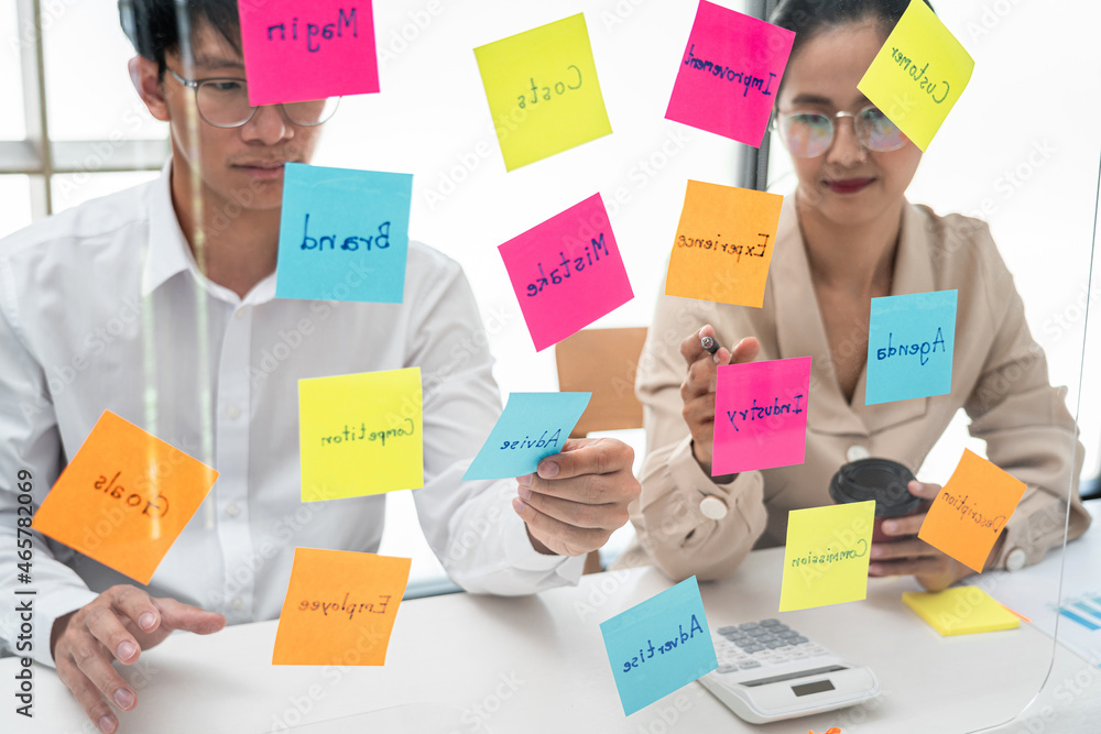 Poster two business people holding and pointing keyword on post it notes on a glass wall to brainstorm abou