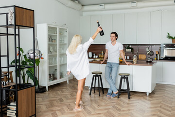 cheerful man looking at blonde woman with bottle of wine and glasses in kitchen