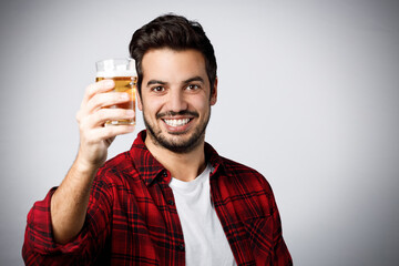 Handsome millennial guy in red plaid shirt smiling and holding a half-pint of beer isolated on grey background.