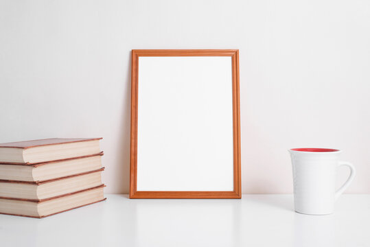 Portrait vertical blank frame mock up. Books, cup of coffee or tea and white picture template with empty space for text or branding standing on table indoors. Home decor with layout elements