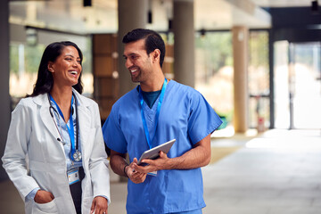 Two Medical Staff In White Coats And Scrubs With Digital Tablet Having Informal Meeting In Hospital