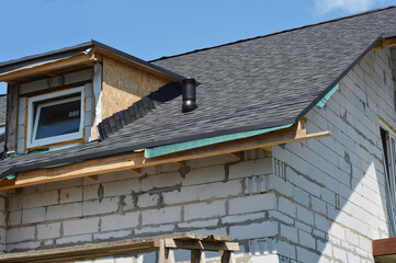 A close-up of a brick house corner under construction with a roof covered with a vapor barrier, asphalt shingles, using flashing near the attic window and roofing ventilation, with incomplete eaves.