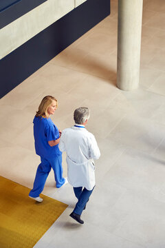 Overhead Shot Of Male And Female Doctors In White Coats And Scrubs Walking Through Busy Hospital