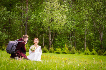 Naklejka na ściany i meble Young photographer take photo of bride sitting on grassy area outdoors with forest panorama