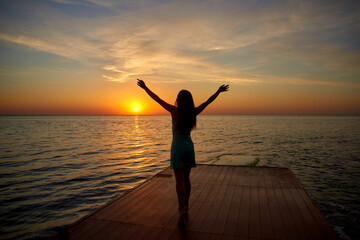 silhouette girl standing on pier raising her hands to sun. sunrise or dawn on sea or ocean side....