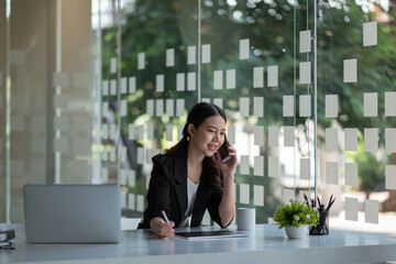 Smiling young asian businesswoman sit at desk smiles talking on smartphone looking out the window enjoy pleasant conversation with client, female sales manager makes successful profitable deal concept