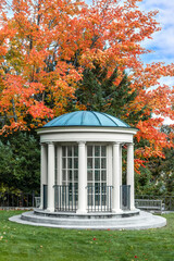 The public library in Camden, Maine is an architectural and landscaping masterpiece with this white, gazebo structure adorning the grounds backed by a tree displaying colorful fall foliage. - 465791441