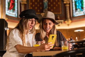 Two tourist friends looking on the phone at leisure activities in a modern cafeteria. Sipping an orange juice, lifestyle