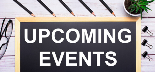 On a light wooden table - a board with the text UPCOMING EVENTS, pencils, plants, glasses and paper clips. Business concept