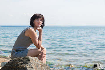 A brunette woman in a striped summer dress sits on a rock against the background of the sea.