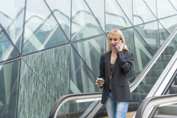 Entrepreneurial girl in business park in a modern building with escalator, young blonde in a suit making a call with a take away coffee