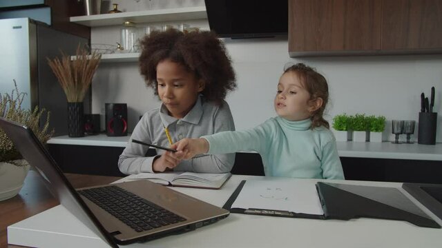 Positive happy adorable multiracial diverse elementary age girls studying online educational lesson using laptop pc, learning to draw pictures while relaxing together in domestic kitchen.
